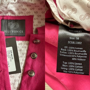 May include: A pink dirndl dress with a black label that says "Juli von CS" and the size "38". The label also lists the fabric composition of the dress, including linen, cotton, and polyester.