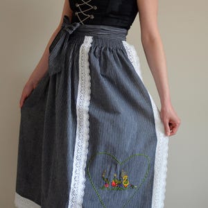 May include: A black and white striped dirndl skirt with white lace trim and an embroidered heart with a scene of people playing music.