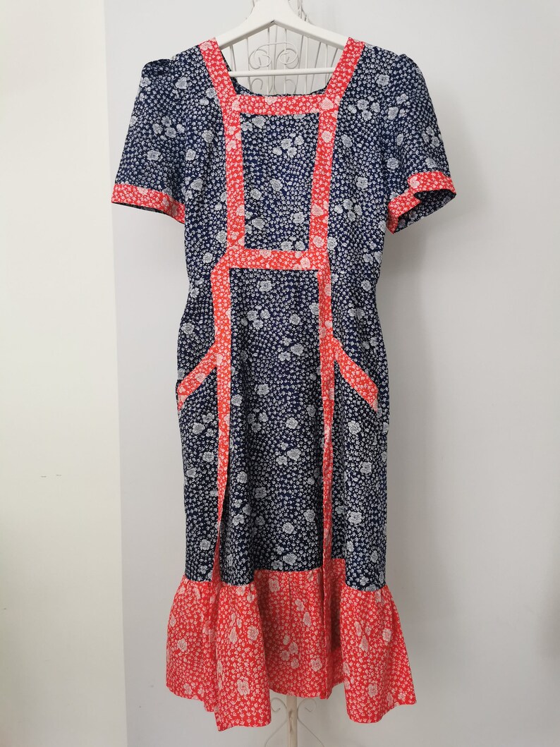 May include: A vintage blue and white floral dress with red trim. The dress has a square neckline, short sleeves, and a full skirt. It is a midi length dress.