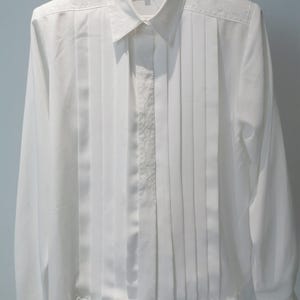 May include: White long-sleeve button-up shirt with a pleated front and embroidered details. The shirt has a collar and a label that reads "Verable".