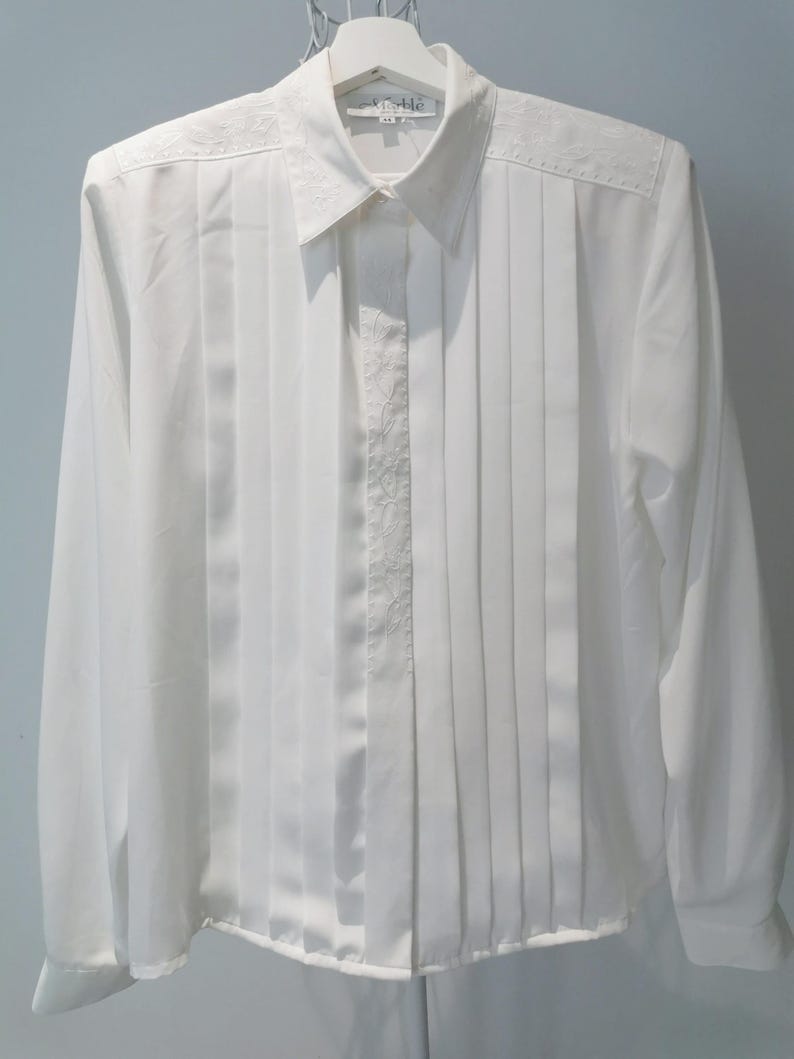 May include: White long-sleeve button-up shirt with a pleated front and embroidered details. The shirt has a collar and a label that reads "Verable".