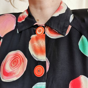 May include: Black button-up shirt with a colorful abstract print of circles and swirls. The shirt has a collar and two orange buttons.