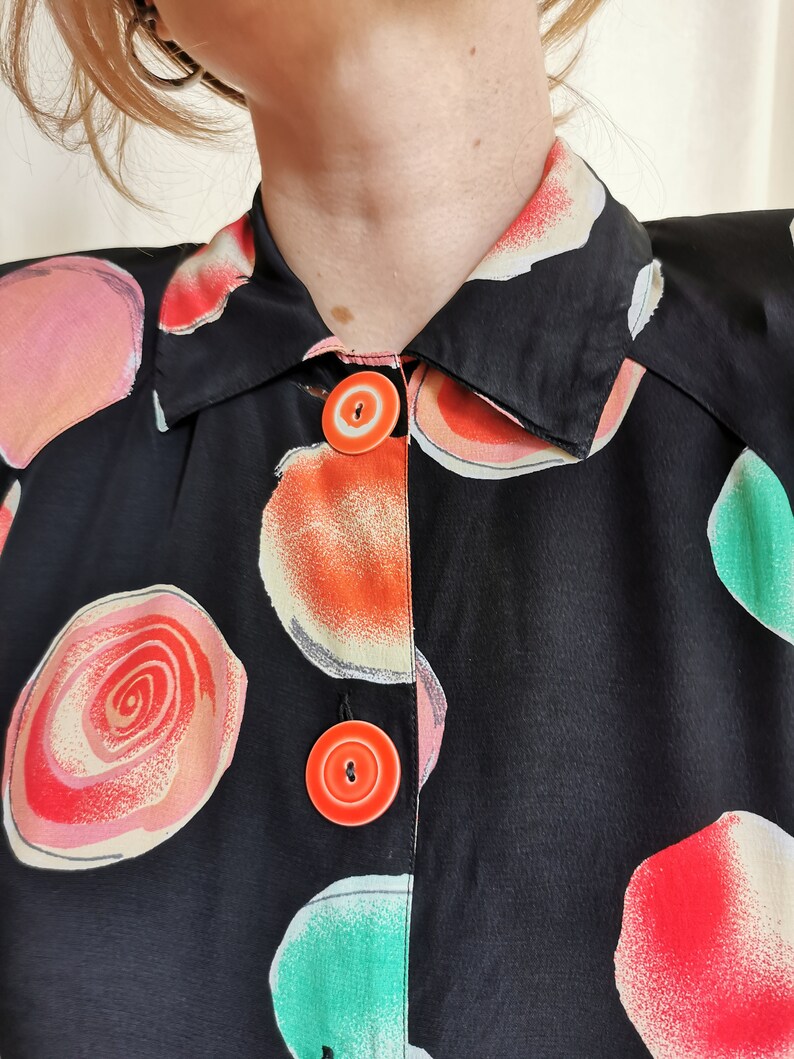 May include: Black button-up shirt with a colorful abstract print of circles and swirls. The shirt has a collar and two orange buttons.