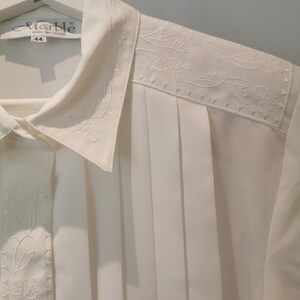 May include: White button-down shirt with a pleated front and embroidered floral details. The shirt has a collar and a label that reads "Marble" with the size "44" printed on it.