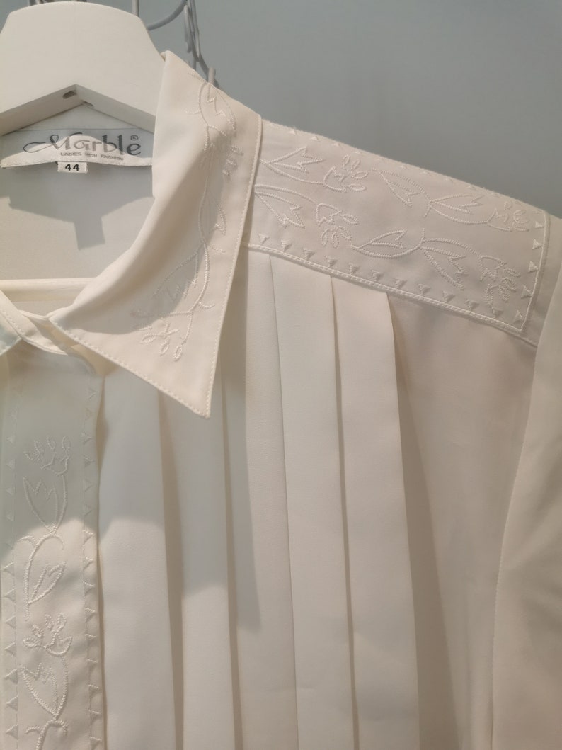 May include: White button-down shirt with a pleated front and embroidered floral details. The shirt has a collar and a label that reads "Marble" with the size "44" printed on it.