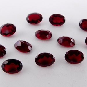 Può includere: Una collezione di gemme ovali di colore rosso intenso. Le superfici sfaccettate delle pietre riflettono la luce, creando un effetto scintillante. Le gemme sono sparse su una superficie bianca, mostrando il loro colore ricco e la loro chiarezza. Adatto per la creazione di gioielli.