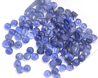 Puede incluir: Una colección de cabujones redondos de piedras preciosas azules. Los cabujones son lisos y pulidos, con una forma ligeramente abovedada. Están dispersos sobre una superficie blanca.