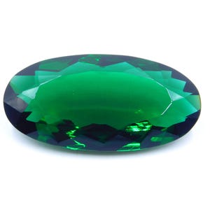 May include: An oval-shaped, faceted gemstone with a deep emerald green color. The gem has a polished surface that reflects light, creating a brilliant shine. The gemstone is set against a white background.