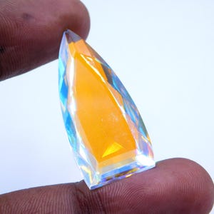 以下が含まれることがあります： 鮮やかなオレンジ色のコアと虹色の多色ファセットを持つ、ファセット加工された細長い宝石。 宝石は2本の指の間に挟まれ、研磨された反射面と鋭い角度が強調されています。 宝石の形は凧に似ています。