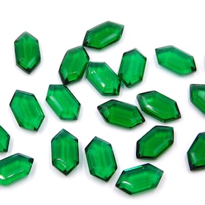 Puede incluir: Una colección de 18 gemas de vidrio facetado de color verde esmeralda, cada una con forma de hexágono con extremos puntiagudos. Las gemas están dispersas sobre un fondo blanco.