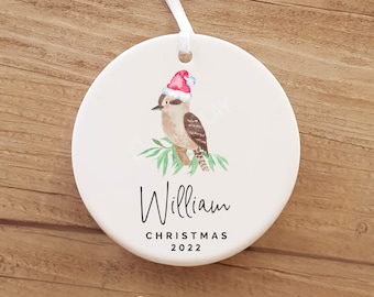 Op de afbeelding: Witte keramische kerstversiering met een aquarel-illustratie van een kookaburra met een kerstmanmuts. De versiering is gepersonaliseerd met de naam "William" en de tekst "Christmas 2022".