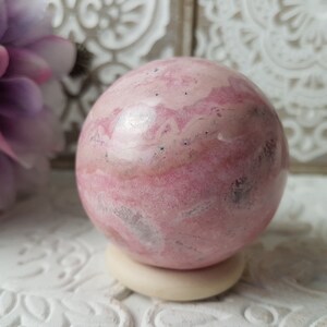 Peut inclure: Une sphère de cristal rose et blanche, posée sur un petit socle blanc cassé. Le cristal présente un aspect marbré avec des nuances de rose et de blanc. En arrière-plan, une fleur violette floue et des éléments décoratifs blancs.