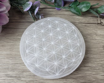 May include: A round, white selenite crystal charging plate with a detailed Flower of Life pattern etched into the surface. The plate is smooth and sits on a wooden surface, with greenery in the background.