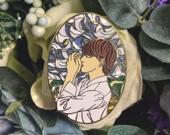 Puede incluir: Un pin de esmalte dorado con una persona de cabello castaño y una camisa blanca, sobre un fondo de vitral azul y verde.