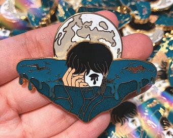 Puede incluir: Un pin de esmalte dorado que representa a una persona con cabello negro y una máscara blanca con una sola lágrima. La persona está parcialmente sumergida en un líquido azul y verde con una luna creciente en el fondo.