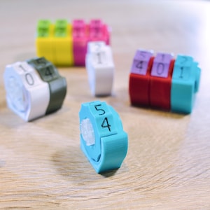 Peut inclure: Une collection de jetons compteurs en plastique multicolores dotés de roues rotatives avec des chiffres noirs imprimés. Les petites unités modulaires sont présentées dans diverses combinaisons de rouge, bleu, vert, blanc, orange et violet.