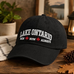 Könnte beinhalten: Eine schwarze Baseballkappe mit weißem Sticktext, der LAKE ONTARIO lautet, gefolgt von dem Text MIN NOW ALWAYS mit zwei roten Ahornblatt-Symbolen zwischen den Wörtern. Die Kappe hat eine strukturierte Krone und einen gebogenen Schirm.