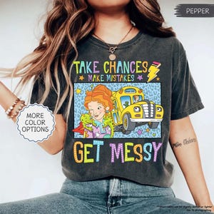 Op de afbeelding: Een antracietgrijs t-shirt met een afbeelding van een vrouw met oranje haar, een groene hagedis en een gele schoolbus, samen met de tekst "TAKE CHANCES MAKE MISTAKES GET MESSY". Het ontwerp bevat een gele bliksemschicht en kleurrijke sterren.