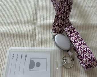 May include: A clear plastic ID badge holder with a white card insert and a retractable lanyard. The lanyard features a burgundy and gold floral pattern. The badge holder has a metal clip and a small metal ring.