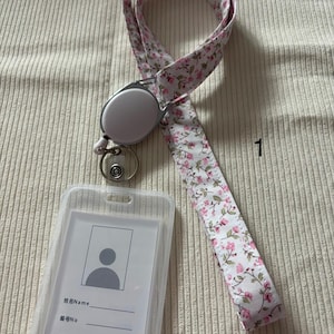 Peut inclure: Un cordon floral avec un enrouleur rétractable et un porte-badge. Le cordon présente un fond blanc avec des fleurs roses et des feuilles vertes. Le porte-badge est en plastique transparent avec une carte d'identité vierge.
