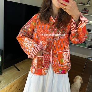 Può includere: Giacca trapuntata arancione con motivi floreali e geometrici, con dettagli a righe rosse e bianche sulle maniche. Il testo "fashionandfianish" è visibile sulla giacca. La giacca è abbinata a una gonna bianca.