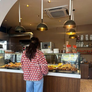 Peut inclure: Une femme portant une chemise à motifs rouge et blanc avec une jupe en jean bleu se tient devant une vitrine de boulangerie. La vitrine est remplie de pâtisseries et de pains. La boulangerie a un intérieur moderne avec des murs blancs et des accents en bois.