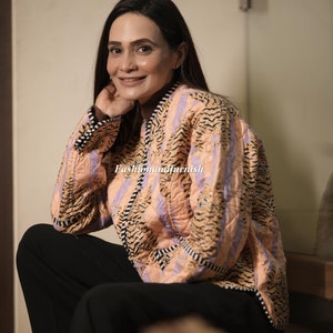 Może przedstawiać: Kobieta w różowej i fioletowej pikowanej kurtce z nadrukiem tygrysa. Kurtka ma czarno-biały pasek. Kobieta siedzi na schodach z kwiatem na pierwszym planie.