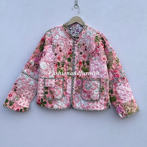 以下が含まれることがあります： ピンク色のキルティングジャケット。花柄の刺繍と白黒のストライプのトリムが施されています。「Fashionandfurnish」の文字が入っています。丸襟、フロントポケット2つ。金属製のハンガーにかかっています。