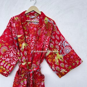 Pode incluir: Robe vermelha com padrão floral e cinto a condizer, pendurada num cabide de madeira. O robe apresenta um design vibrante com elementos florais rosa, verdes e brancos. O texto "Fashionandfurnish" é visível.