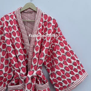 Puede incluir: Bata acolchada con un estampado de fresas rojas sobre fondo blanco. La bata tiene un interior y ribetes a rayas, con cinturón y bolsillos a juego. El texto "Fashionandfurnish" es visible.