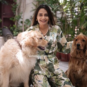 May include: A woman wearing a green and white patterned pyjama set with a white pearl necklace sits on a wooden floor with two dogs. The dogs are a golden retriever and a brown and white cocker spaniel.