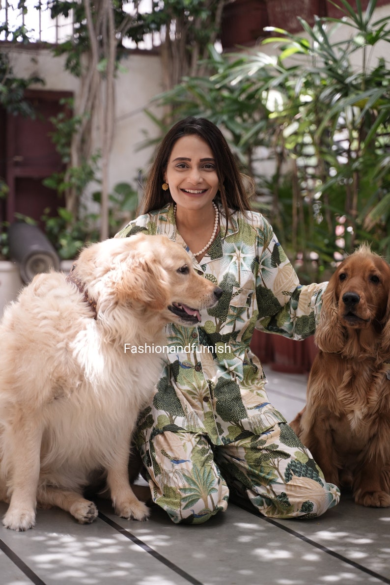 May include: A woman wearing a green and white patterned pyjama set with a white pearl necklace sits on a wooden floor with two dogs. The dogs are a golden retriever and a brown and white cocker spaniel.