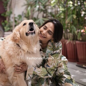 May include: A woman wearing a green and white patterned jumpsuit smiles and hugs a golden retriever dog. The dog is sitting on a grey tiled floor.