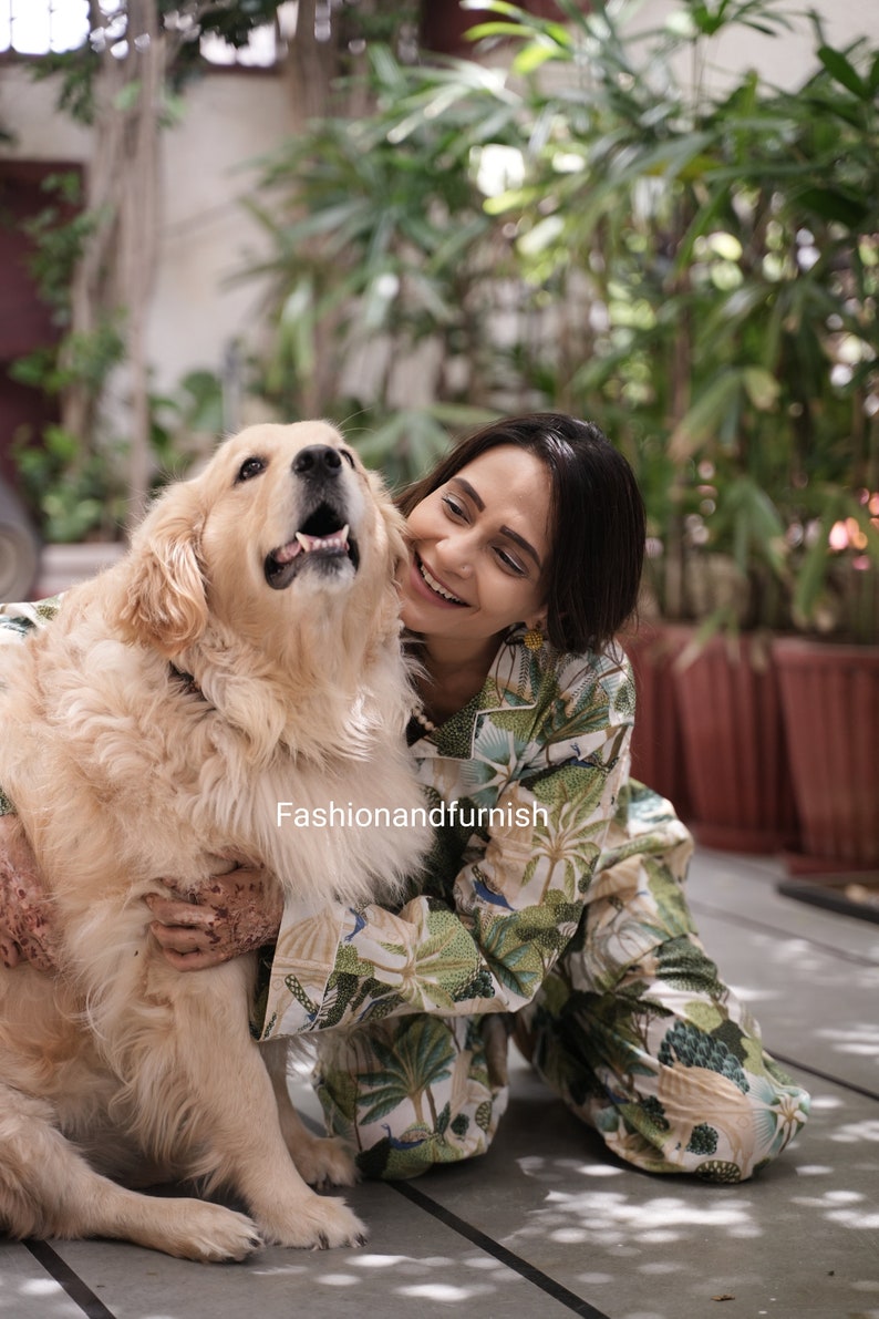 May include: A woman wearing a green and white patterned jumpsuit smiles and hugs a golden retriever dog. The dog is sitting on a grey tiled floor.