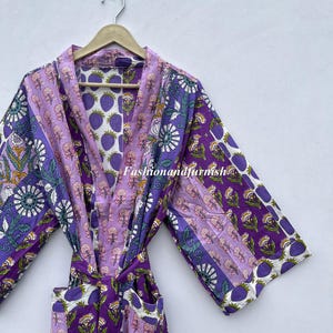 May include: A patterned purple and lavender robe with a matching belt, hanging on a wooden hanger. The robe features floral and fruit designs. The text "Fashionandfurnish" is visible on the robe.