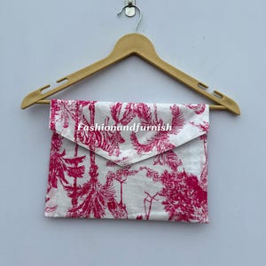 May include: A white and pink patterned clutch with a flap closure hangs from a wooden hanger. The clutch features a botanical design with the text "Fashionandfurnish". The color scheme is white and pink.