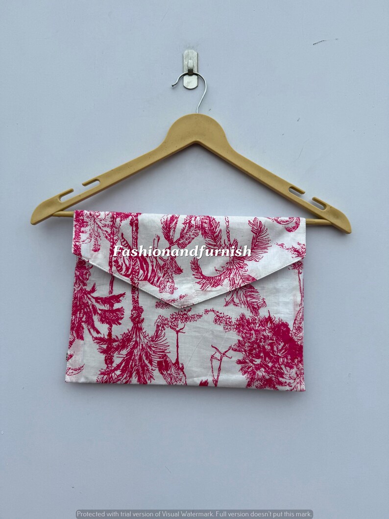 May include: A white and pink patterned clutch with a flap closure hangs from a wooden hanger. The clutch features a botanical design with the text "Fashionandfurnish". The color scheme is white and pink.