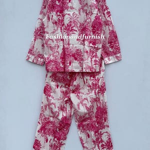 May include: A two-piece pajama set in white and fuchsia. The set includes a long-sleeved button-down shirt and matching pants. The fabric features a repeating pattern of tropical trees, birds, and other foliage. The text "Fashionandfurnish" is visible on the shirt.