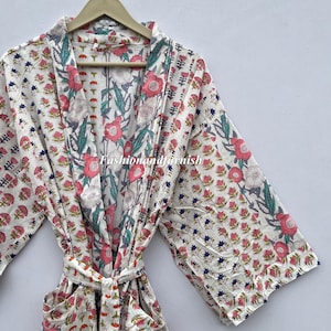 Könnte beinhalten: Weißes Bademantel mit floralem Druck mit rosa und blauen Akzenten. Der Bademantel hat einen Bindegürtel und Taschen. Das Blumenmuster umfasst rosa Blüten, blaue Akzente und grüne Blätter. Der Bademantel hängt an einem Holzbügel.