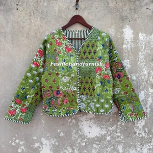 以下が含まれることがあります： 鮮やかなグリーンのベースに、多色の花柄と幾何学模様のブロックプリントが施された長袖のキルティングジャケット。Vネック、フロントボタン留め、黒と白のストライプの縁取りが特徴的なデザインの衣類です。