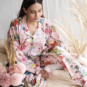 May include: A light pink pajama set featuring a vibrant floral print with red, blue, yellow, and green flowers and leaves. The long-sleeved button-down top has a collar and a chest pocket, paired with matching long pants. The text Fashionandfurnish is visible.