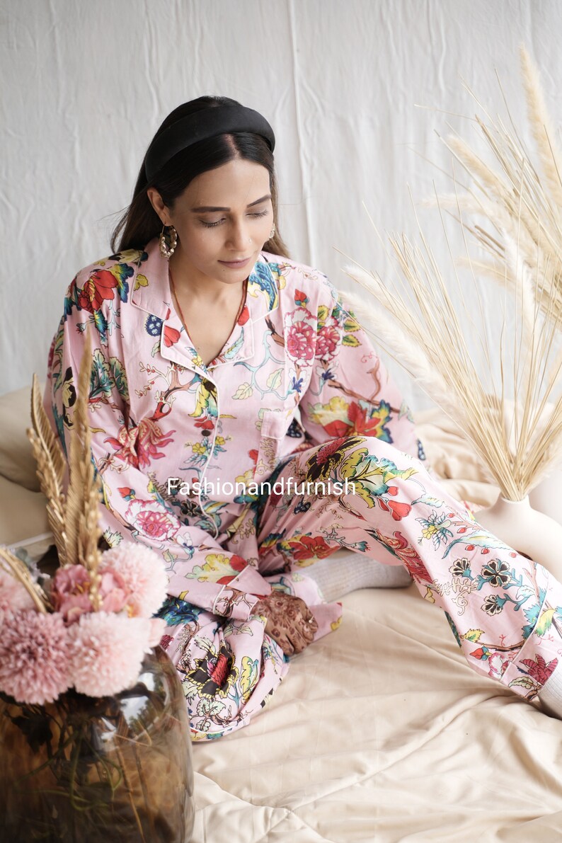 May include: A light pink pajama set featuring a vibrant floral print with red, blue, yellow, and green flowers and leaves. The long-sleeved button-down top has a collar and a chest pocket, paired with matching long pants. The text Fashionandfurnish is visible.