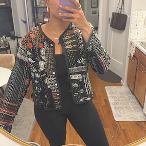 Puede incluir: Una chaqueta acolchada negra con un patrón floral y geométrico. La chaqueta tiene un cierre de botón y mangas largas.