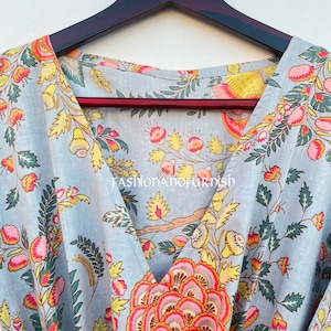 Puede incluir: Prenda azul claro con estampado floral, con flores y hojas rosas, amarillas y verdes. Se aprecia el cuello en V. La prenda est&aacute; colgada en una percha de madera oscura.