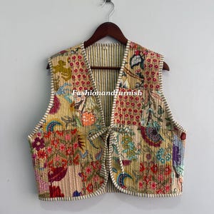 Op de afbeelding: Een gewatteerd vest met een bloemen patchwork ontwerp in tinten geel, roze, rood en blauw. Het vest heeft een striksluiting en is afgewerkt met witte biezen. De tekst "Fashionandfurnish" is zichtbaar.