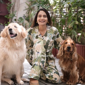 May include: A woman wearing a green and white patterned pyjama set with a white pearl necklace, sitting on a wooden floor with two dogs. The dogs are a golden retriever and a brown cocker spaniel.
