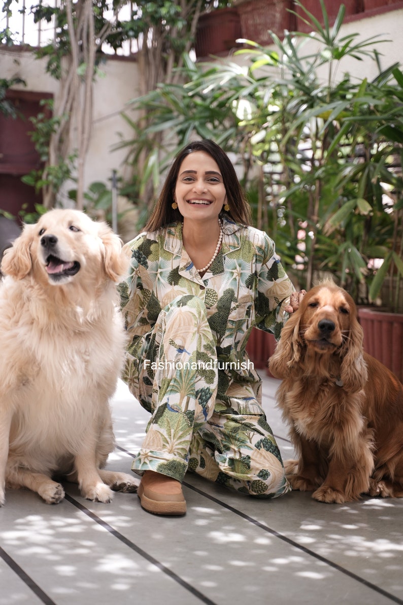 May include: A woman wearing a green and white patterned pyjama set with a white pearl necklace, sitting on a wooden floor with two dogs. The dogs are a golden retriever and a brown cocker spaniel.