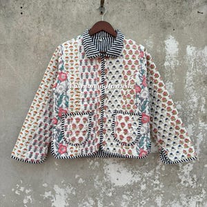 May include: A quilted jacket featuring a white base with a repeating floral pattern in pink, orange, and blue. The garment has a black and white striped collar, cuffs, and pocket trim, with a button-down front and the text Fashionandfurnish.