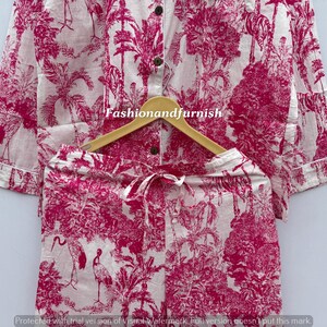 May include: A two-piece pajama set with a white background and a pink toile pattern featuring birds, trees, and foliage. The long-sleeved top has buttons, and the shorts have a drawstring. The set is displayed on a wooden hanger.
