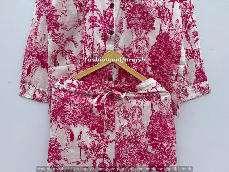 May include: A two-piece pajama set with a white background and a pink toile pattern featuring birds, trees, and foliage. The long-sleeved top has buttons, and the shorts have a drawstring. The set is displayed on a wooden hanger.